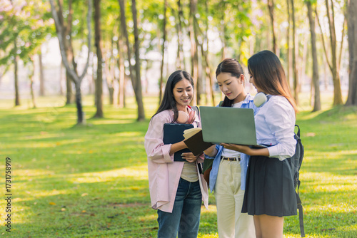Group of Asian college students standing on green grass and discussing with laptop and books. Concept of teamwork, education, and outdoor learning in a natural environment.
