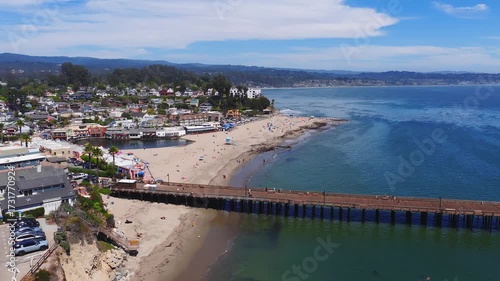 Wallpaper Mural Aerial view of Capitola, California, showing a wooden pier, sandy beach with umbrellas, coastal buildings, and gentle ocean waves under a clear sky. Torontodigital.ca