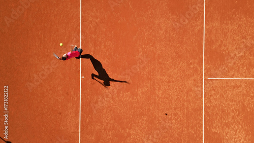Tennis player in red shirt and black shorts stretches dynamically on a clay court, showcasing focus and agility. Aerial shot highlights sports training and skill development in a sunlit setting.