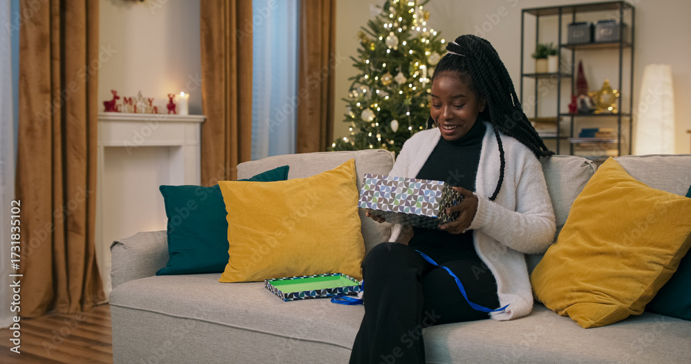 Obraz premium Sitting on a couch, a young student of multi-racial background unpacks a Christmas present, eagerly awaiting a miracle. She opens the box, pulls out a warm black hat, rejoices, and tries it on.