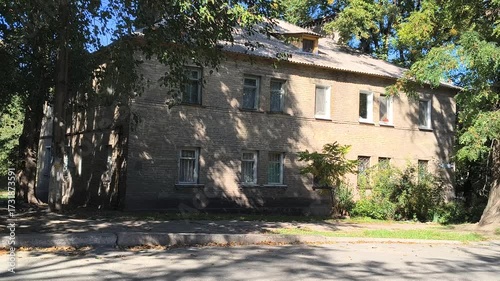  A quiet residential street with a two-story brick house shaded by tall trees. Sunlight filters through leaves, casting soft shadows. A peaceful urban scene with vintage charm and greenery.