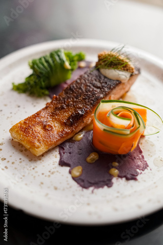 A gourmet dish with crispy-skinned salmon fillet, served with purple yam puree, carrot and cucumber ribbons, green vegetable, and a dollop of cream topped with fresh herbs.