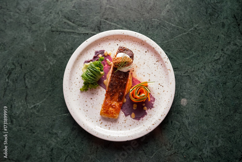 A gourmet dish with crispy-skinned salmon fillet, served with purple yam puree, carrot and cucumber ribbons, green vegetable, and a dollop of cream topped with fresh herbs.