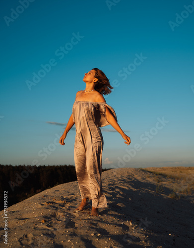 A girl in a gray dress walks on a sand dune at sunset in summer