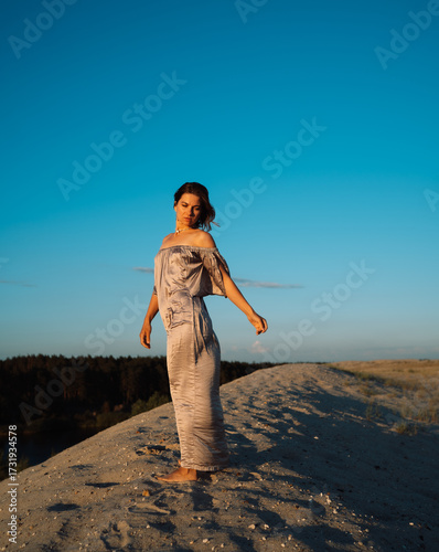 A girl in a gray dress walks on a sand dune at sunset in summer