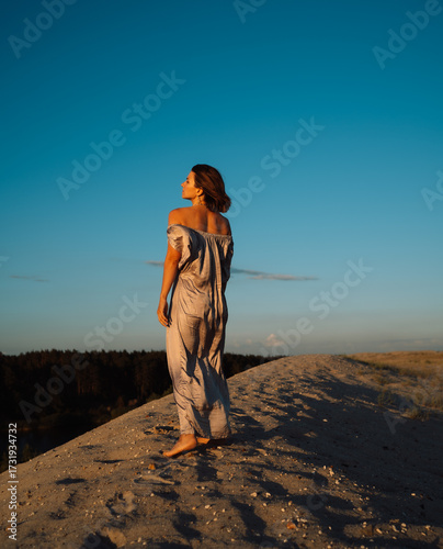 A girl in a gray dress walks on a sand dune at sunset in summer