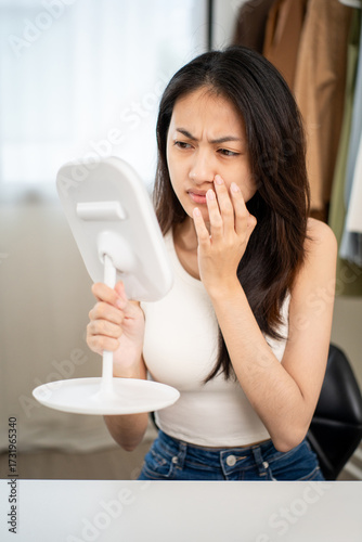 Worried Asian woman looking at mirror, touching dry irritated skin and acne spots. Female feeling unhappy with wrinkles and face dehydration searching for skincare solution.
