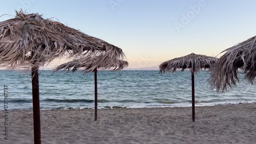 Scenic beach view with straw parasols and calm ocean waves at sunset. The sandy shore and rustic umbrellas create a relaxing summer holiday atmosphere, perfect for themes of travel, vacation, leisure
