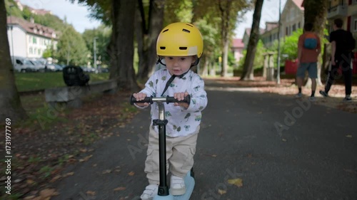 Wallpaper Mural Toddler riding scooter on tree-lined park path wearing yellow helmet, focused and playful, enjoying carefree summer day while older sibling walks behind Torontodigital.ca
