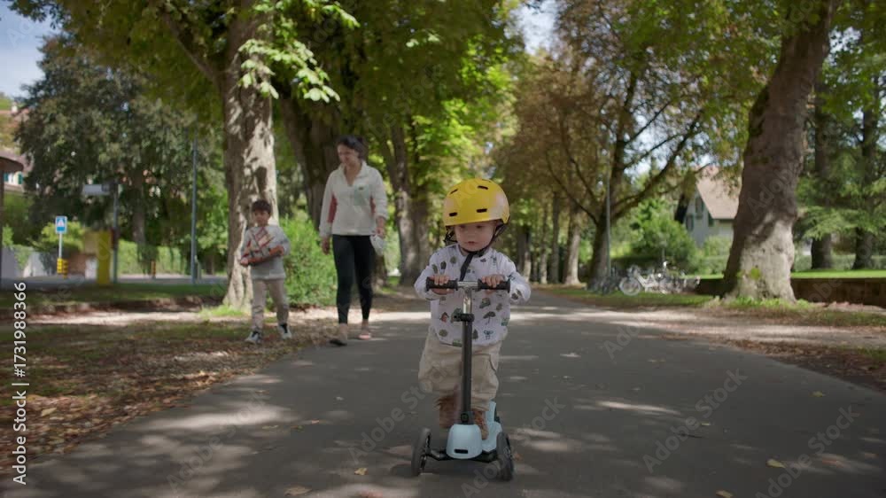 custom made wallpaper toronto digitalToddler riding scooter on pathway wearing yellow helmet, mother and older sibling walking behind, family enjoying time together outdoors in peaceful park setting