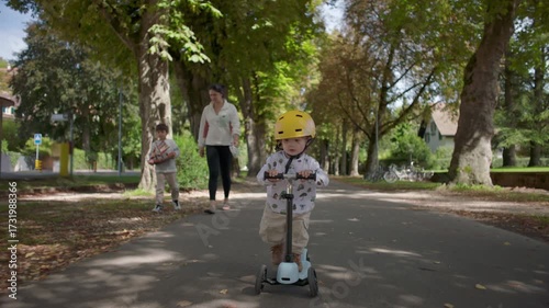 Wallpaper Mural Toddler riding scooter on pathway wearing yellow helmet, mother and older sibling walking behind, family enjoying time together outdoors in peaceful park setting Torontodigital.ca