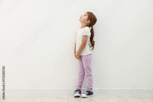 Cute little girl standing near white wall and looking up. Space for text