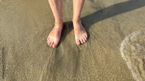 A close-up of bare feet walking along a wet sandy beach near the shoreline, with gentle waves washing over nearby rocks. The sunlight casts a soft shadow, enhancing the natural textures of sand, water