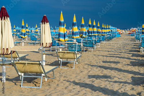 Sun loungers on sandy beach with Umbrellas. Empty seaside resort. Italy. Coastline row chaise lounge sunbathe. Blue rainy sky in summer