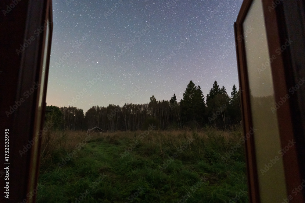Fototapeta premium A tranquil view from an open door, overlooking a grassy field and the starry night sky in rural Estonia.