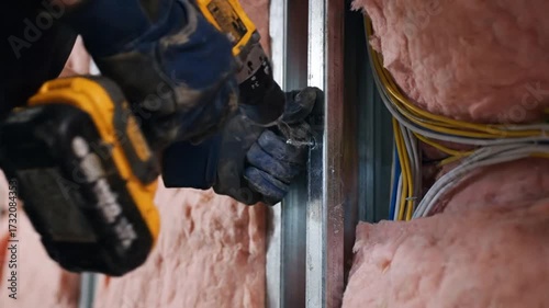 A construction worker in gloves uses a cordless power drill to fasten a screw into a metal stud with pink insulation.