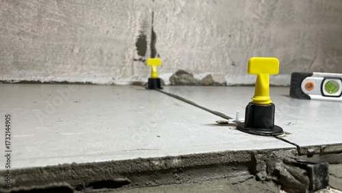 Close-up of ceramic tile leveling system with yellow wedges and black clips on unfinished floor during installation process, ensuring flat alignment and even grout joints in construction.
