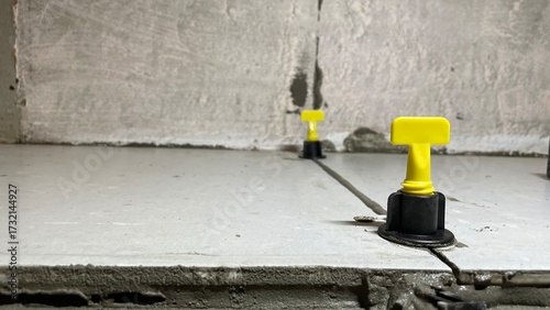 Close-up of ceramic tile leveling system with yellow wedges and black clips on unfinished floor during installation process, ensuring flat alignment and even grout joints in construction.