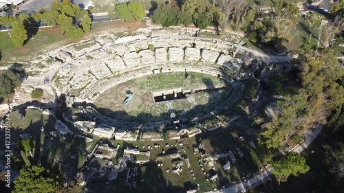 Cinematic 4K drone footage of the Roman Amphitheater in the Neapolis Archaeological Park, Syracuse, Sicily, filmed on a bright summer day with stunning views of the ancient ruins and greenery