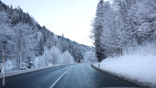 Snowy scenic mountain icy road near Hinterglemm surrounded by conifer forest early morning twilight. Cars with headlights drive along icy asphalt, creating a typical alpine winter travel scene