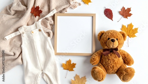 Autumn baby clothes flat lay with teddy bear, knitted blanket, frame, brown overalls, and yellow leaves on beige background.