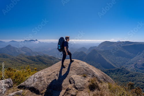 A hiker with a backpack on the mountaintop along the long-distance trail between the cities of Petrópolis and Teresópolis, in the mountainous region of Rio de Janeiro, enjoying a beautiful panoramic v