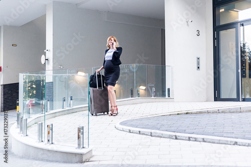 Pregnant businesswoman walking with luggage and talking on phone