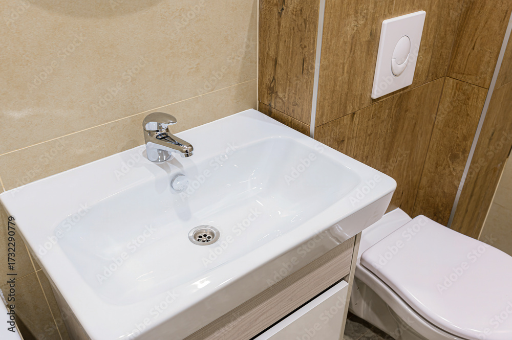 Fototapeta premium Close-up shot of a modern bathroom featuring a white sink with a chrome faucet, and a toilet. Tiled walls in neutral tones and wood paneling enhance the clean, contemporary design