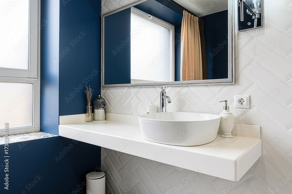 Fototapeta premium A chic bathroom featuring a modern, round white sink, a clean white countertop, a framed mirror, and stylish white herringbone tiles against a deep blue wall