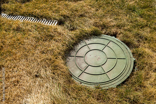 Overhead view of a green septic tank lid nestled in dry, golden grass next to a white drainage grate