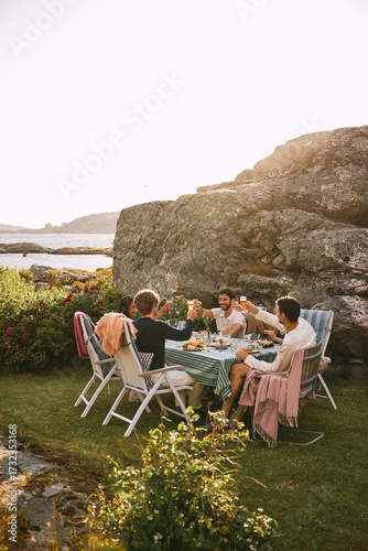 Group of male and female multiracial friends doing celebratory toast while sitting near hill for dinner party under clea