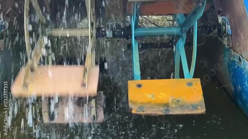 Rotating catamaran paddles in water, showcasing mechanical movement and splashes. Wooden and metal paddle wheels powered by pedal drive, propulsion system. Energy production.