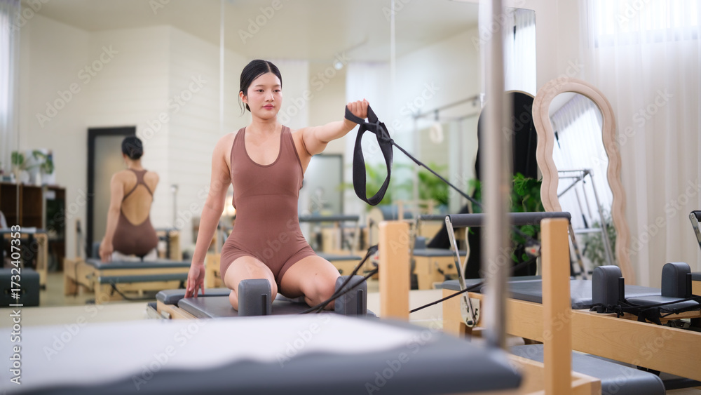 Fototapeta premium Woman kneeling on Pilates reformer, holding resistance strap to begin workout.