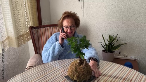 Elderly woman sitting at home talking on phone while touching plant on table calmly