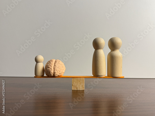 A seesaw balances a small peg figure and a brain model against two larger peg figures, all on a dark wooden surface with a white background.