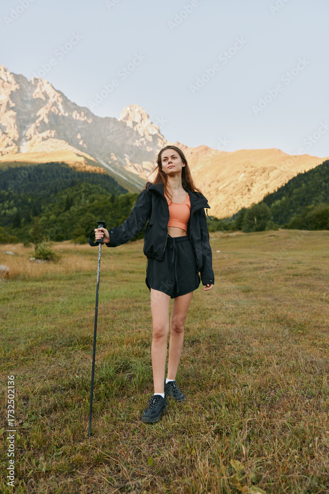 Fototapeta premium A confident outdoor enthusiast stands in a grassy alpine meadow with a trekking pole, wearing a jacket, shorts, and boots. Sunlit mountains rise behind, creating a vivid adventure scene.