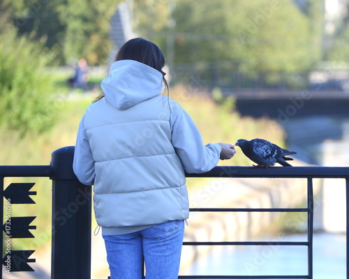 A young woman in a white jacket is feeding a pigeon near a fence