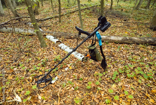 A metal detector is positioned on the ground in a forest, partially covered by colorful autumn leaves. Sunlight filters through the trees, creating a serene atmosphere