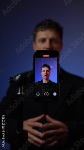 Young man in suit giving energetic motivational speech hitting palm with fist smartphone recording blue background, vertical video