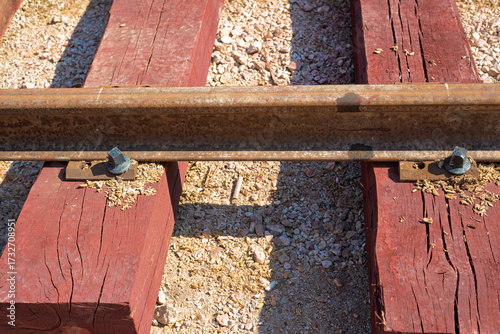 Extreme close-up of a rusted steel rail fastened to a red-brown wooden sleeper, showing the texture of the wood, metal, and light-colored gravel ballast.