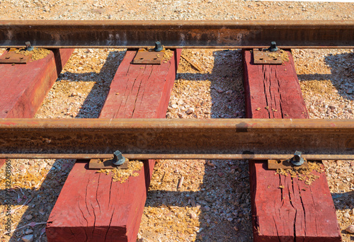 Close-up view of weathered steel railway rails secured to vibrant red-brown treated wooden sleepers, set on a bed of light gravel ballast.