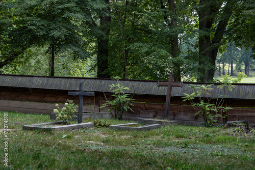 This peaceful image depicts a quaint rural cemetery featuring several weathered wooden crosses standing in a lush green grassy field. Surrounded by dense forest trees and small wooden structures 