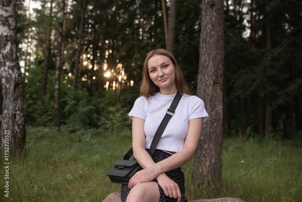 Obraz premium Young woman with long hair wearing a white shirt and black skirt stands confidently in a lush green forest, showcasing a serene outdoor lifestyle and natural beauty