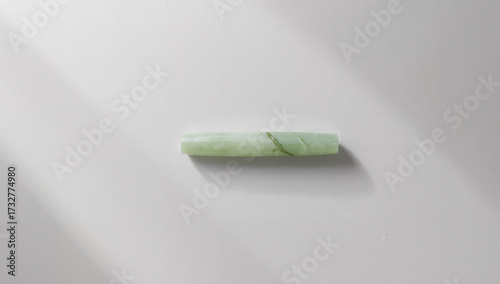 Minimalist flatlay of a single oblong jade stone with natural veins on a clean white background, featuring a diagonal shadow for a modern skincare and wellness aesthetic.