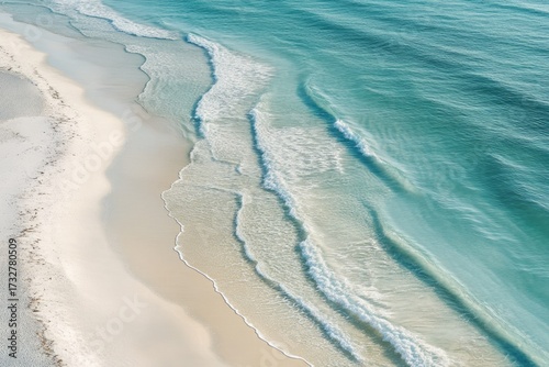 An aerial view captures the serene beauty of a pristine white sand beach meeting the vibrant turquoise ocean. Gentle waves elegantly roll onto the shore, creating a tranquil coastal scene.