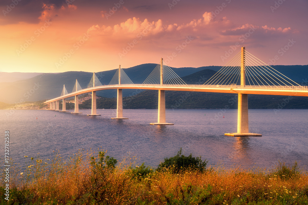 Obraz premium Modern Peljesac bridge and blue sea at colorful sunset in summer in Croatia. Beautiful Peljeski bridge during golden hour, mountains, reflection in water and orange sky with clouds. Architecture