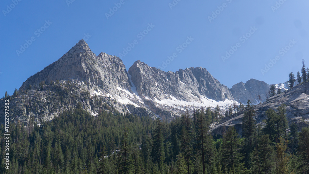 Obraz premium View of jagged mountain peaks with patches of snow and a forest under a clear blue sky day light
