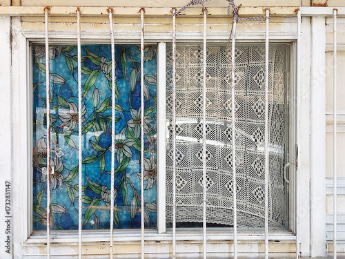 the left with vibrant floral stained glass, the right with a sheer crochet curtain. Rusty metal bars and a weathered wall frame the window, blending bright patterns with aged, neglected textures.