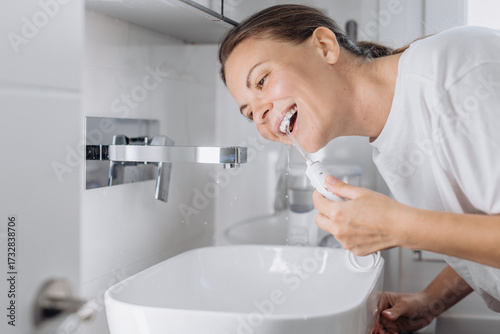 A young woman uses a water flosser to clean her teeth in the bathroom in the morning. Concept: morning routine and oral hygiene at home.