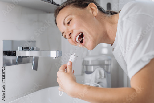 A young woman uses a water flosser to clean her teeth in the bathroom in the morning. Concept: morning routine and oral hygiene at home.